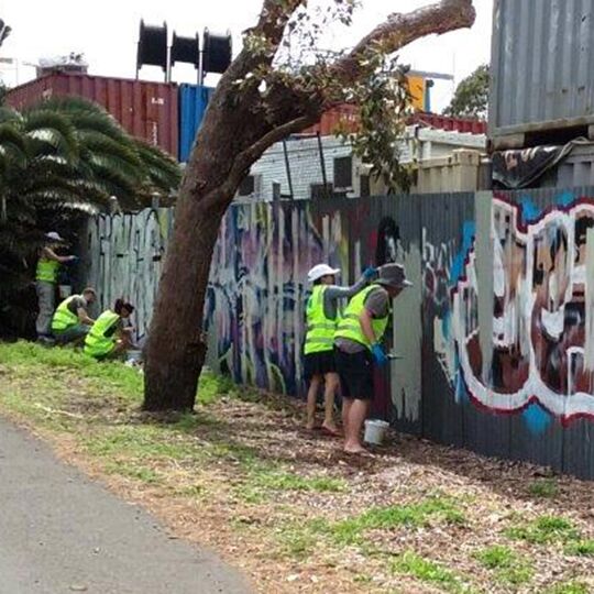  Five council workers in fluorescent yellow vests cleaning graffiti off corrugated iron fences
